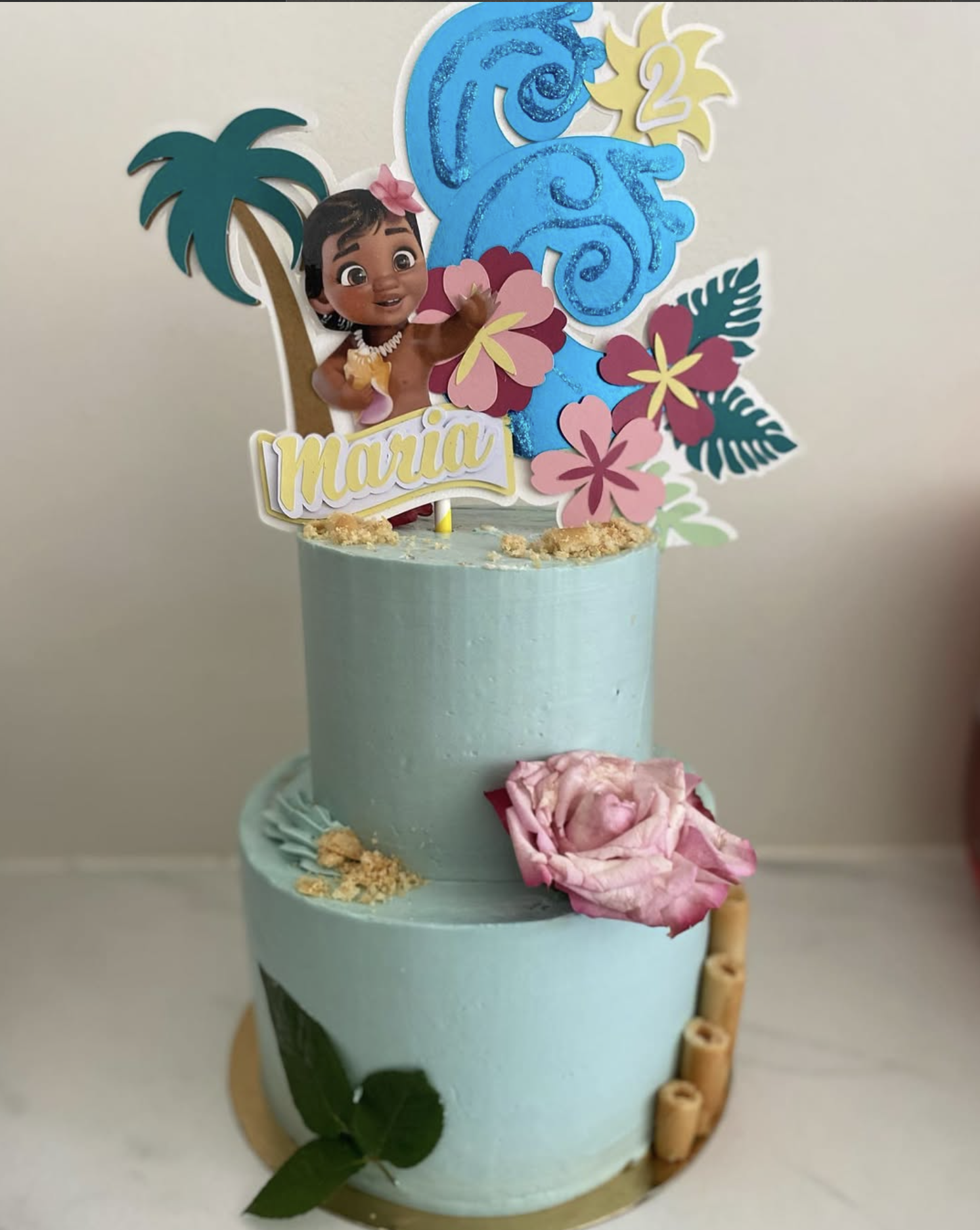 Gâteau anniversaire tropical | Taasty Cakes Gâteau anniversaire tropical — Une création à deux étages aux tons bleu et vert, décorée de fleurs tropicales, d’un personnage et d’un âge personnalisé | Taasty Cakes