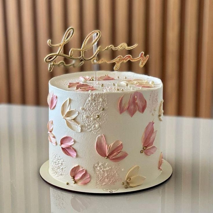 Gâteau floral personnalisé | Taasty Cakes Gâteau floral personnalisé — Un élégant gâteau blanc décoré de fleurs délicates aux tons rose poudré, de détails dorés et d’un topper personnalisé. U | Taasty Cakes