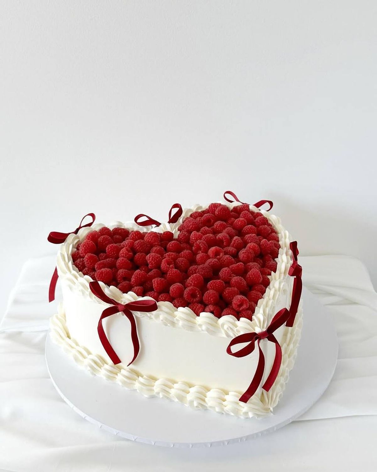 Mini gâteau cœur framboise | Taasty Cakes Mini gâteau cœur framboise — Un petit gâteau cœur délicatement décoré de crème pochée, de framboises fraîches et de rubans rouges. | Taasty Cakes